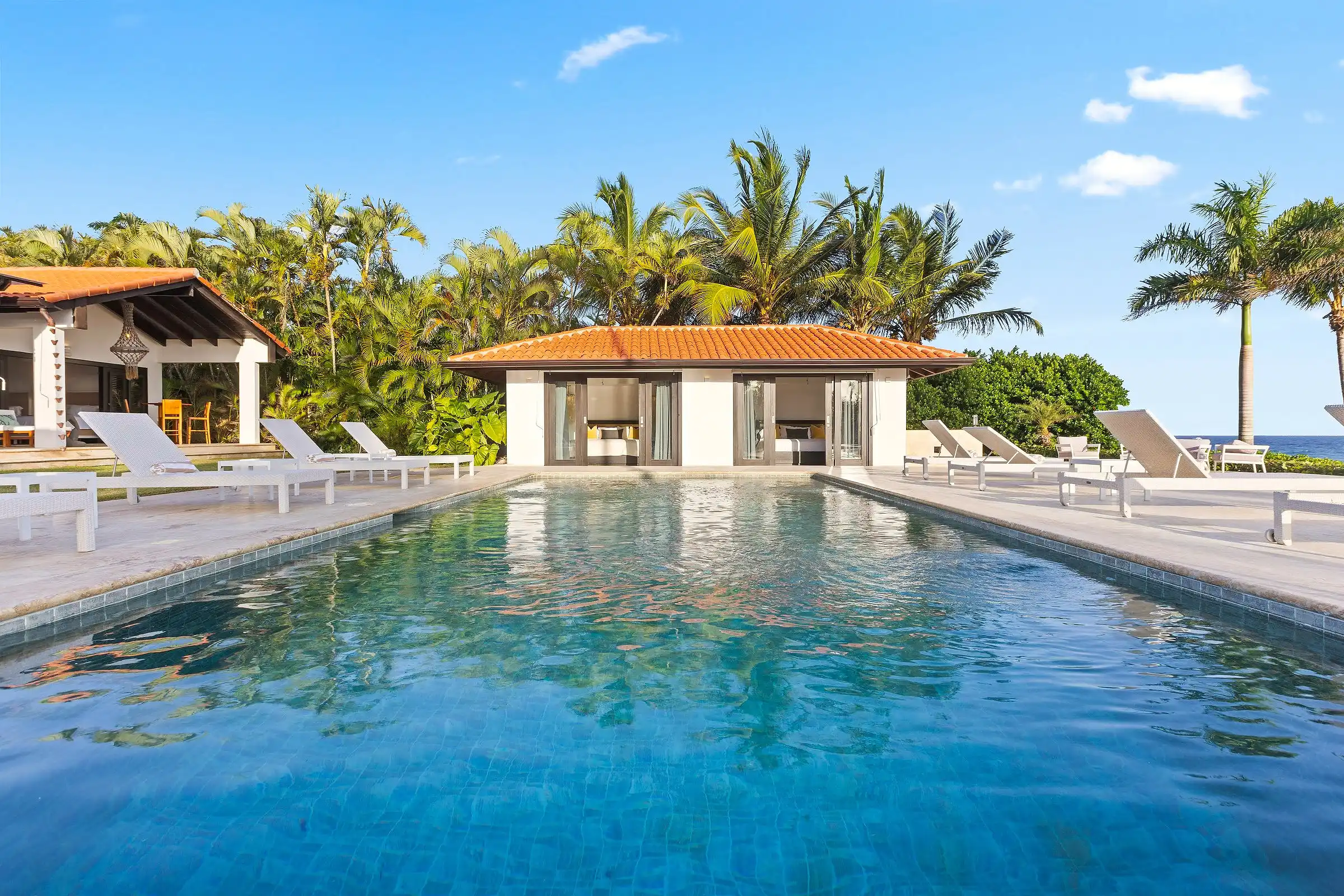 The full length of the pool at Costa Verde 3 framed by tall palms, with the poolhouse and the sea behind